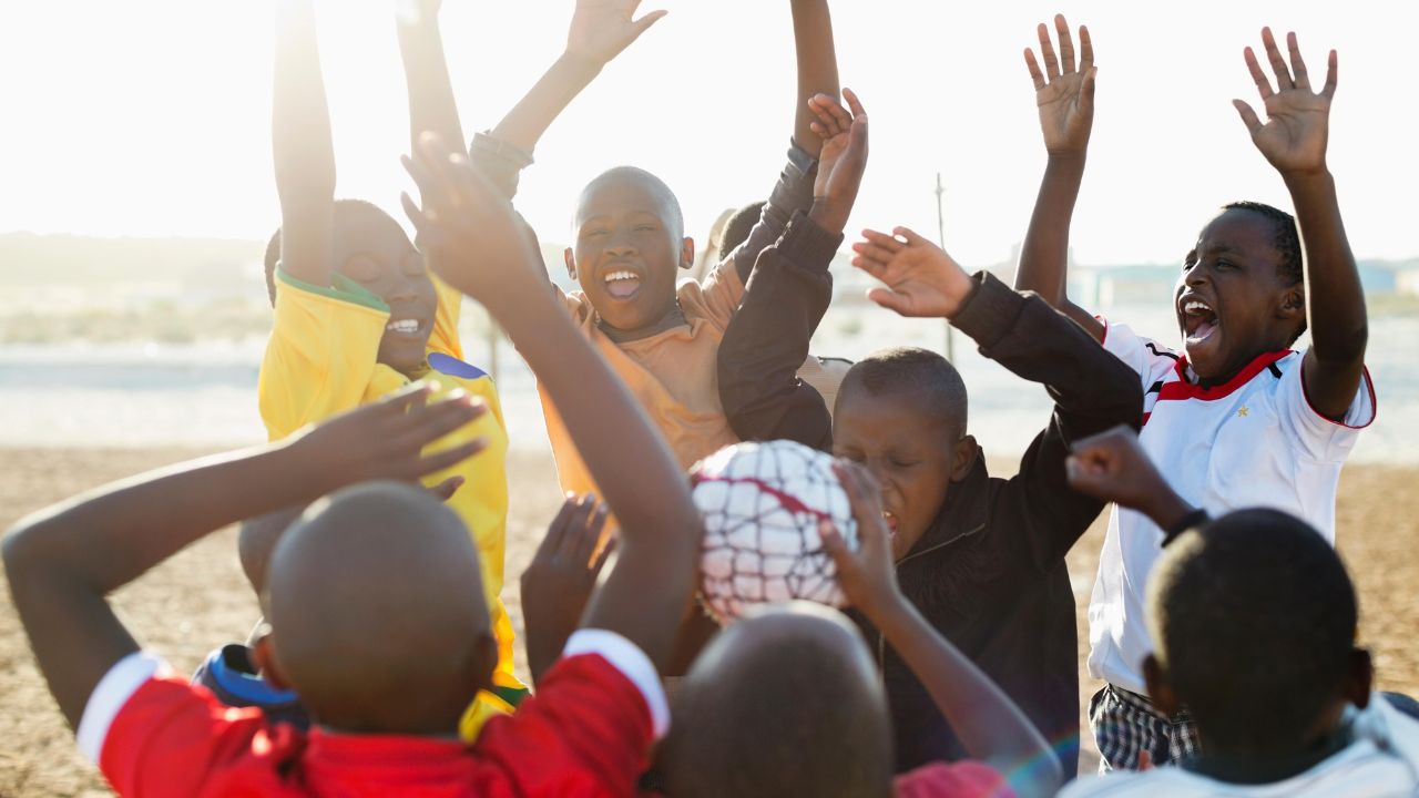 A close-up, sunny outdoor shot of a diverse group of African children, wearing soccer shirts, celebrating with great excitement. They have their arms raised high in the air and are cheering loudly, surrounding a soccer ball held near the center of the group.